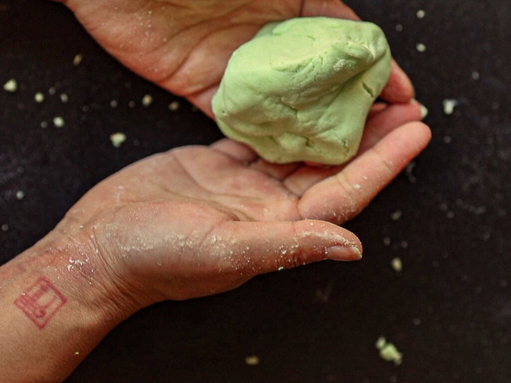 Hands holding a lump of green dough over a dark surface, with a small amount of dough crumbs scattered around. A tattoo is visible on one wrist.